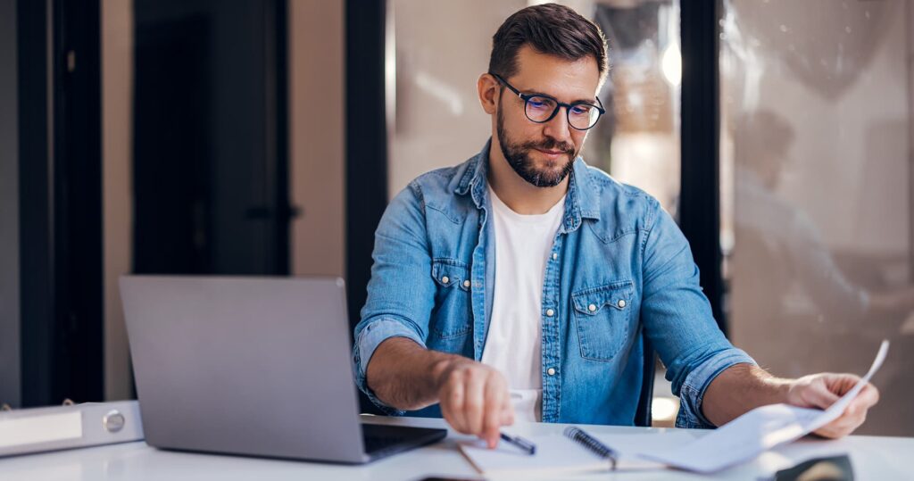 Man in glasses sits behind laptop with papers