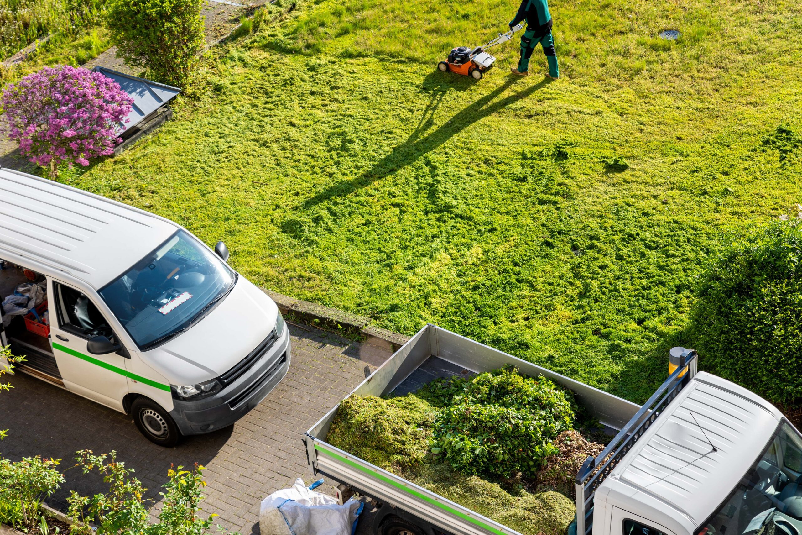 A lawn care company employee mowing a lawn and company vehicles parked at a jobsite.