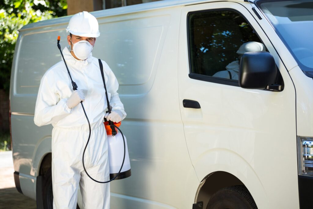 A pest control in a white uniform holding a canister with spray for pests standing next to a work van.